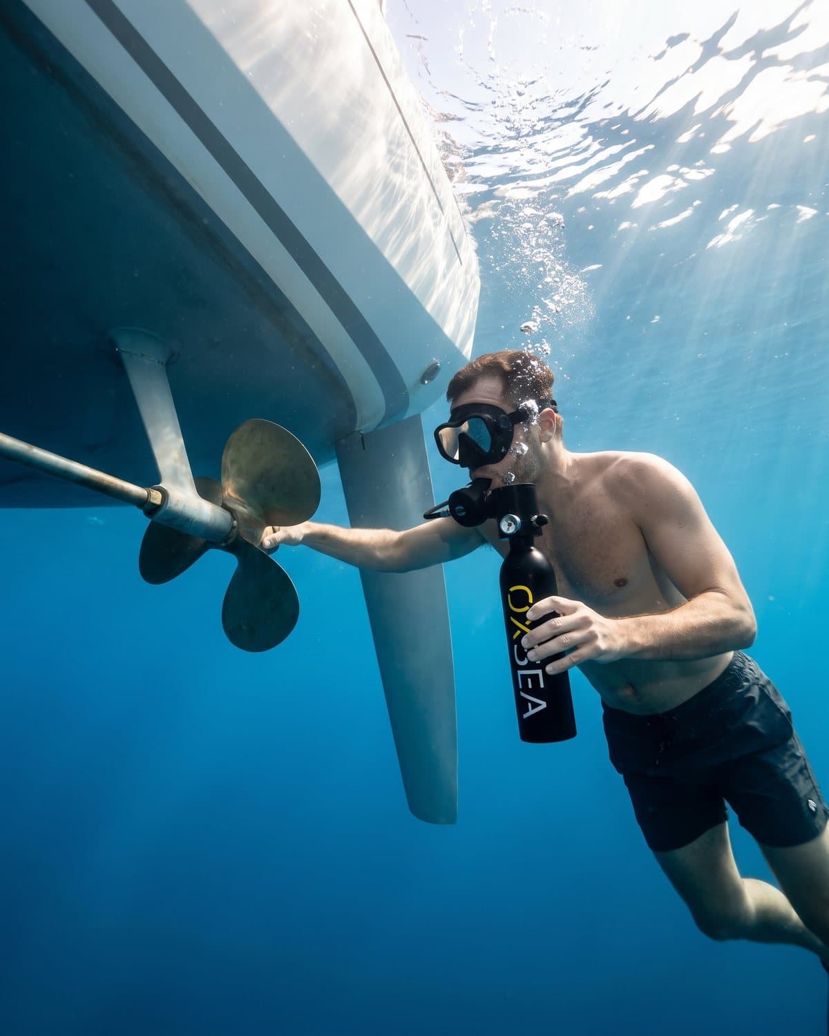 Nageur sous la coque d'un voilier, une main sur l'hélice, la bouteille OXSEA en bouche